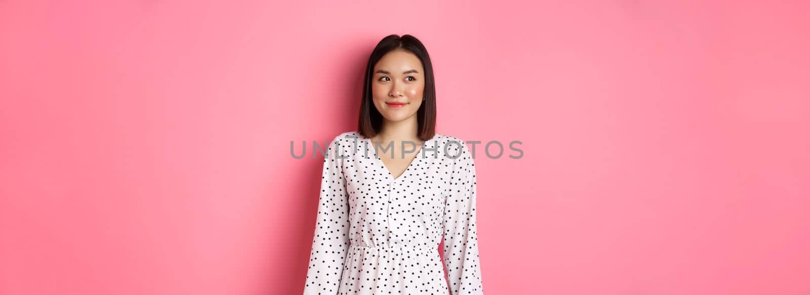 Cute asian woman smiling, looking left at copy space, standing on romantic pink background.