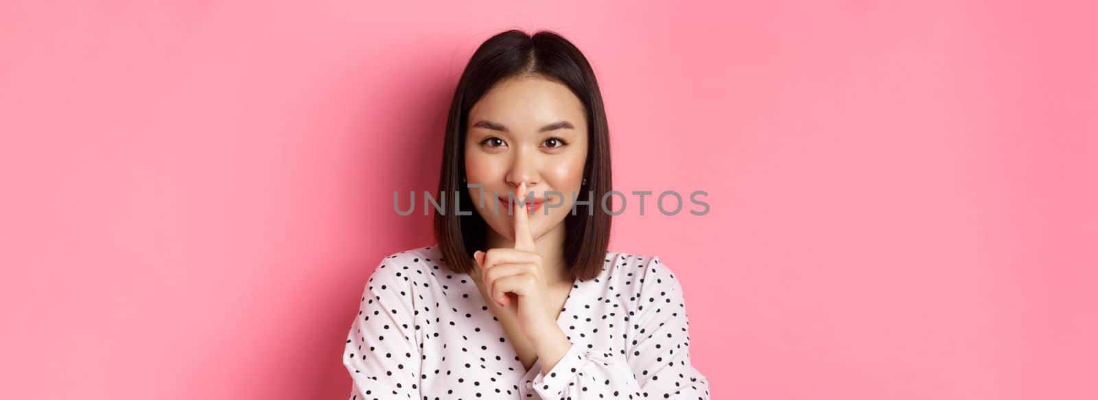 Close-up of mysterious asian woman hiding a secret, hushing and telling to keep quiet, standing over pink background.