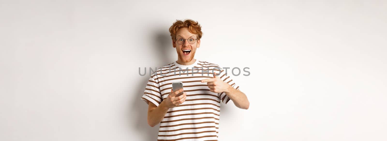 Technology and e-commerce concept. Amazed redhead man checking out online promo offer on mobile phone, pointing finger at smartphone and smiling, white background.