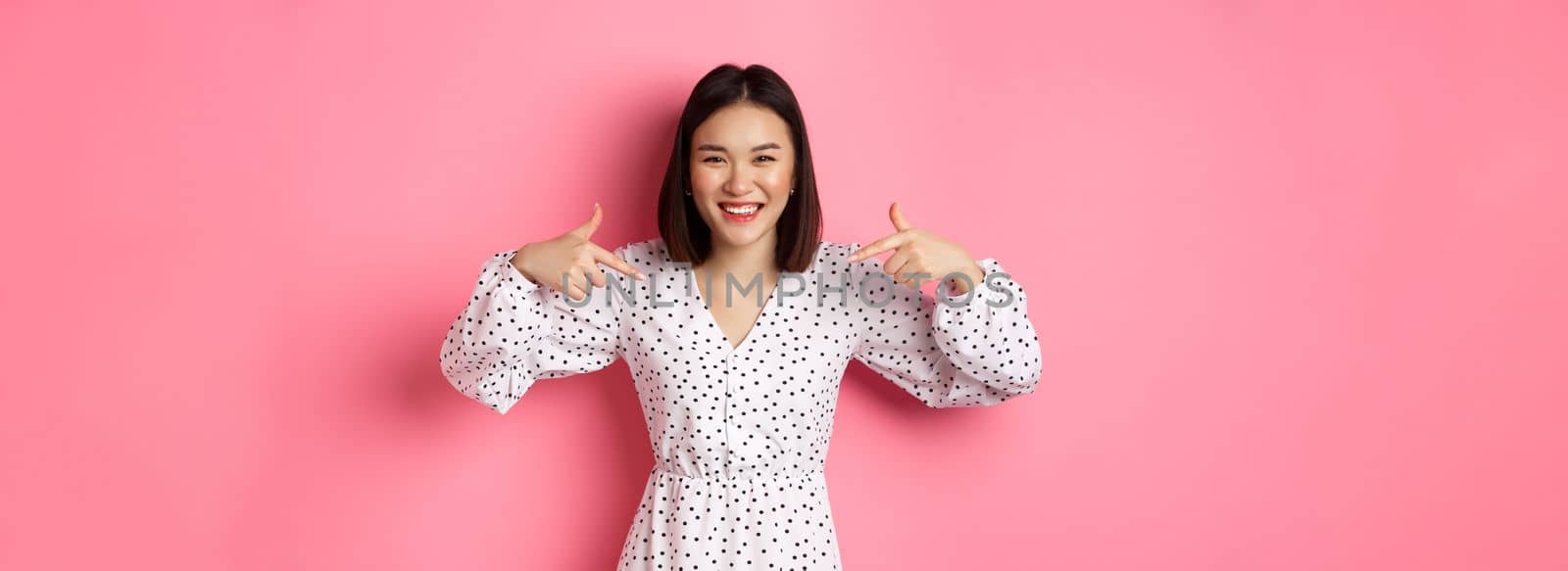 Beautiful korean woman pointing fingers at your logo and smiling, standing in dress over romantic pink background. Copy space