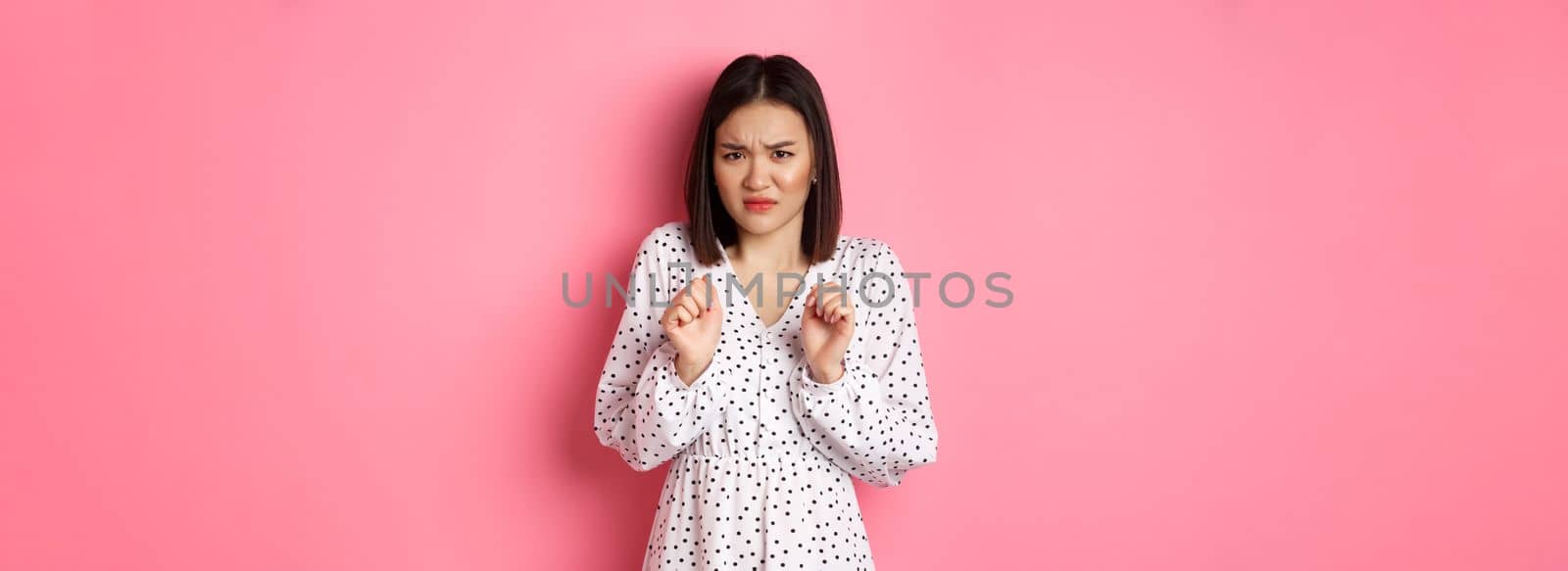 Disgusted asian woman staring with aversion and dislike, frowning and grimacing dissatisfied, standing in dress against pink background.