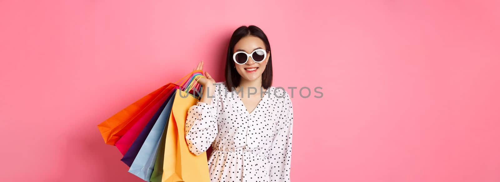 Young asian woman in sunglasses going shopping, holding bags from malls and stores and smiling, standing over pink background.