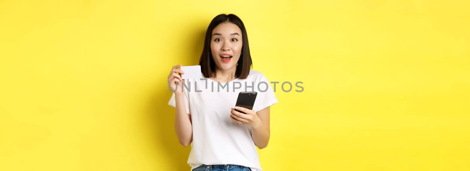 E-commerce and online shopping concept. Excited asian woman order in internet, holding smartphone and plastic credit card, standing over yellow background by Benzoix