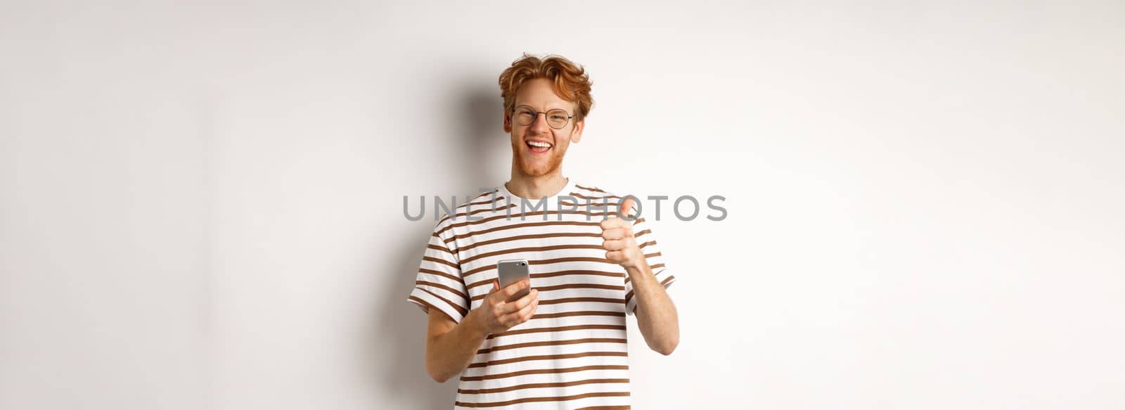 Technology and e-commerce concept. Satisfied male model with red hair, showing thumbs-up and holding smartphone, white background by Benzoix