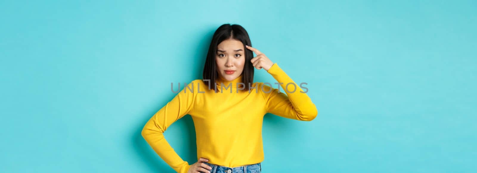 Are you stupid. Portrait of skeptical asian woman scolding someone crazy or dumb, pointing finger at head and staring at camera with dismay, blue background.