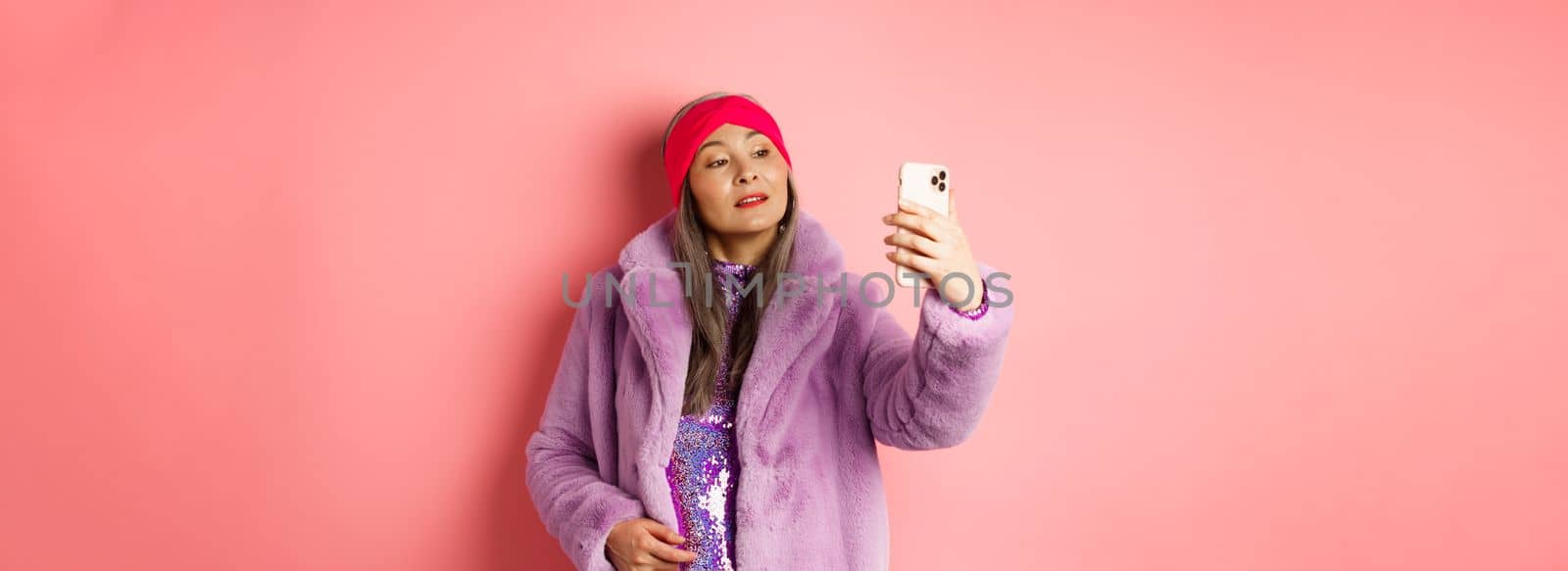 Fashion concept. Stylish asian senior female taking selfie on smartphone, posing in purple faux fur coat and party dress, standing over pink background.