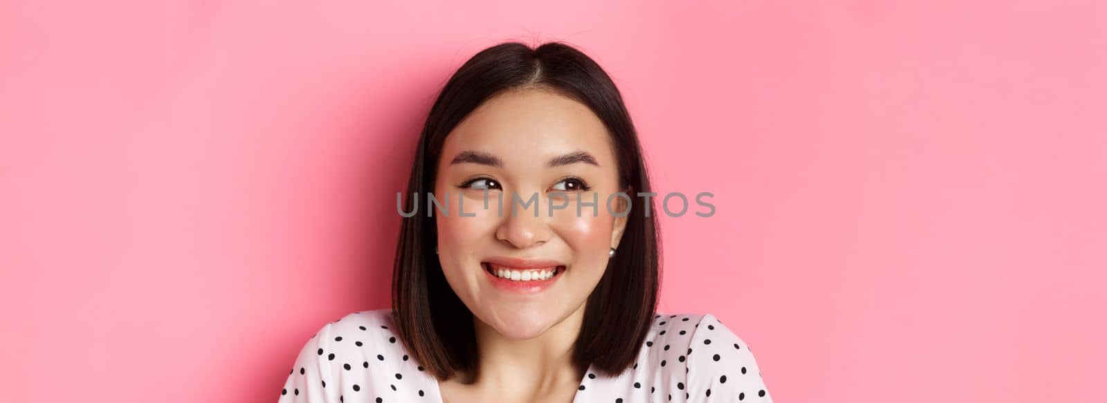 Beauty and lifestyle concept. Headshot of excited asian girl smiling, looking left at promo banner, standing over pink background. Copy space