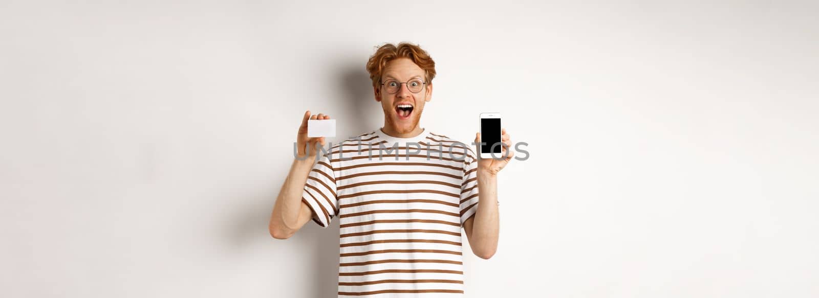 Shopping and finance concept. Young man showing blank mobile screen and plastic credit card, smiling at camera, white background by Benzoix