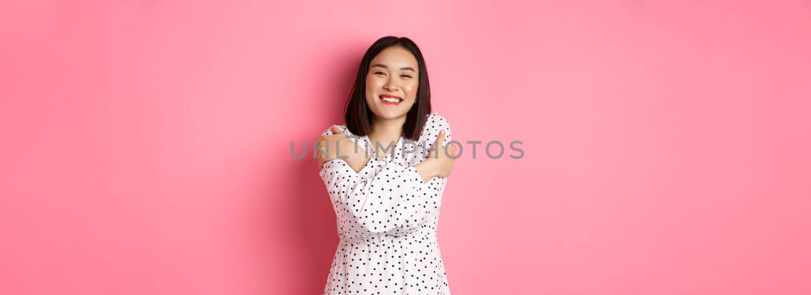 Beautiful asian girl embracing own body, hugging herself and smiling with happy face, looking at camera carefree, standing over pink background.