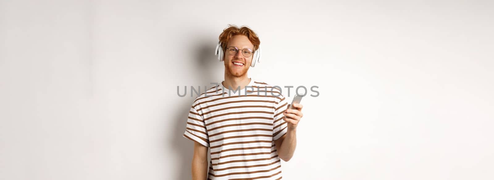 Technology concept. Young man with red hair and beard listening music in headphones and using smartphone, smiling at camera, white background.