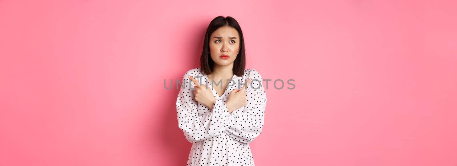 Concerned asian woman having doubts, pointing sideways and looking left with hesitant and sad face, need help with choice, standing over pink background.