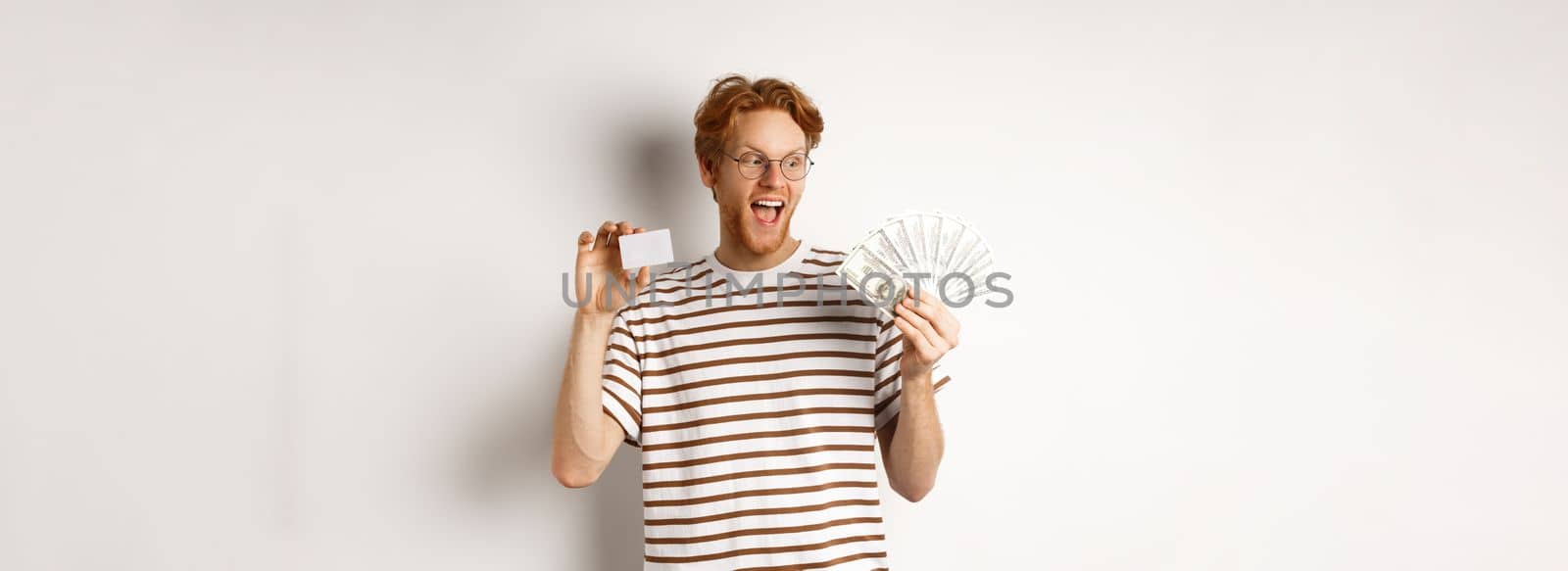 Shopping and finance concept. Cheerful handsome redhead man showing plastic credit card and cash, smiling amazed, white background.