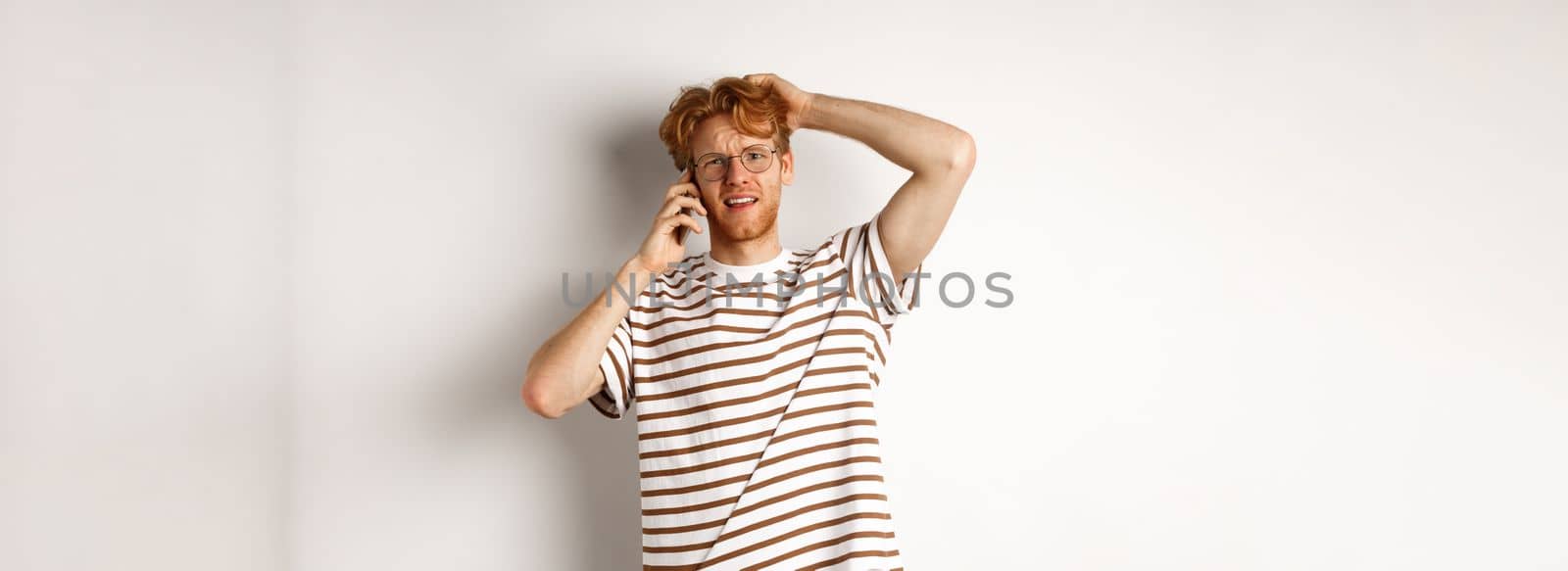 Puzzled redhead guy talking on phone, scratching head and looking confused or indecisive, standing over white background.