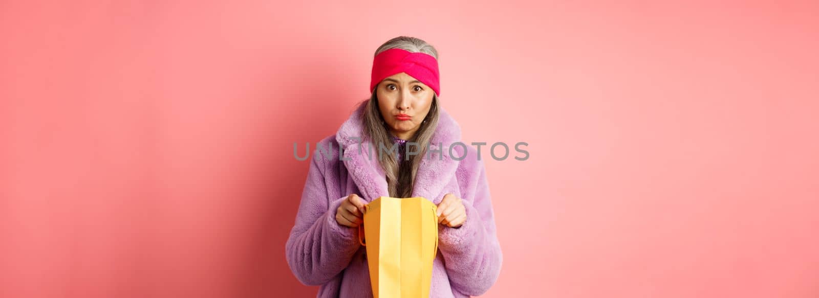 Upset senior asian woman open shopping bag and looking sad at camera, standing over pink background.