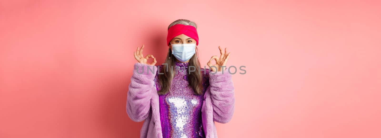 Covid-19, social distancing and fashion concept. Fashionable asian senior woman in face mask and glitter dress, showing okay signs in approval, pink background.