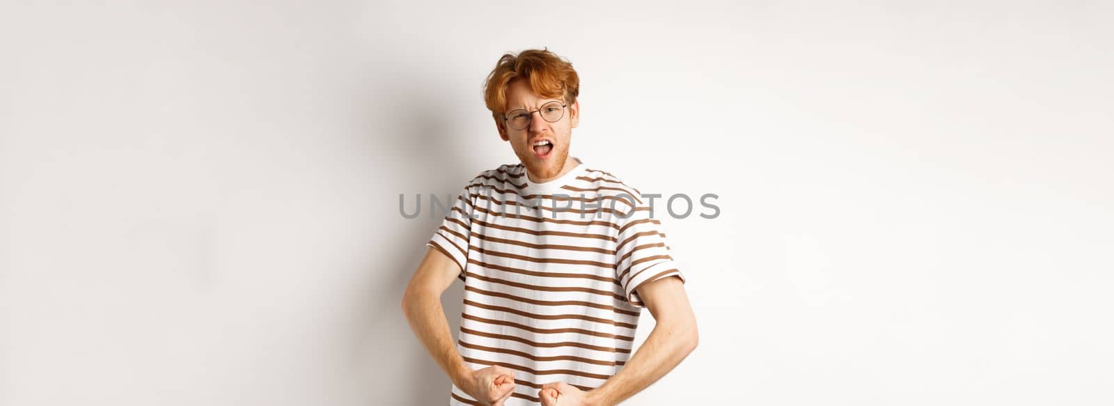 Image of confident and strong redhead man flexing biceps, showing muscles after gym, standing over white background.