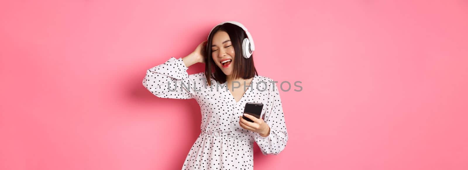 Carefree asian woman having fun, dancing and listening music in headphones, holding mobile phone, standing in dress over pink background.