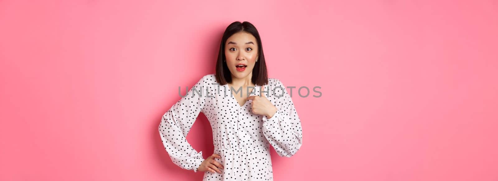 Beautiful brunette asian woman pointing finger at camera, looking surprised as being chosen, standing in dress against pink background.