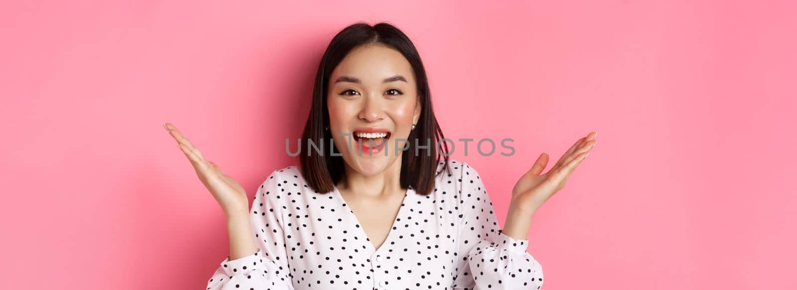 Close-up of surprised and happy asian woman rejoicing, spread hands sideways and smiling, hear great news, standing over pink background.