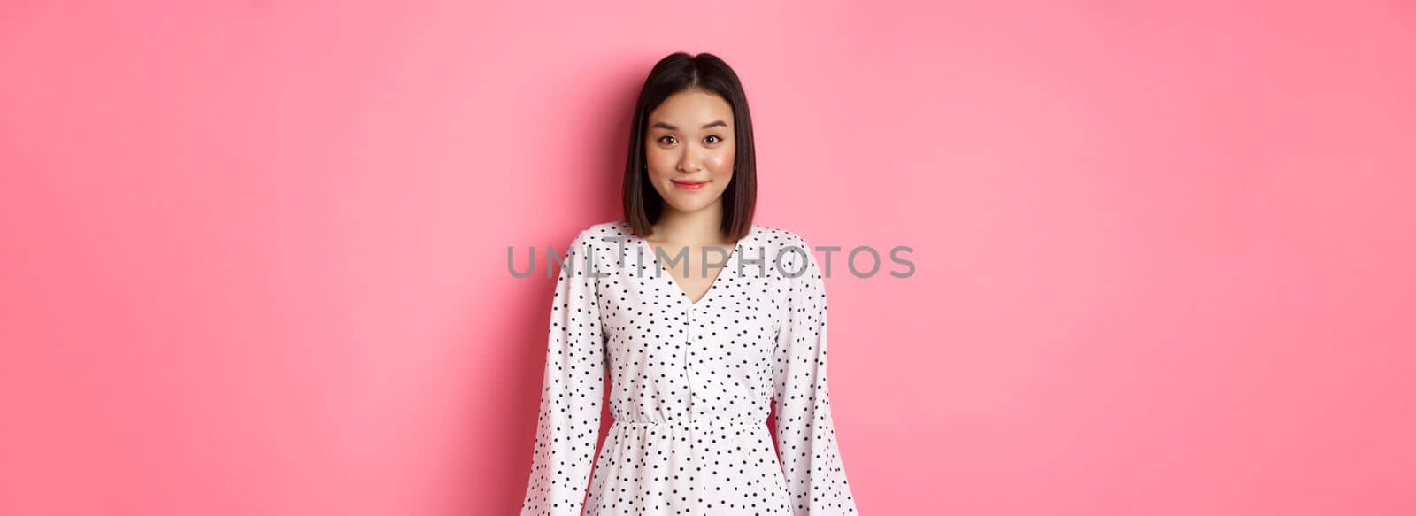 Beautiful asian lady smiling at camera, wearing cute romantic dress, standing over pink background. Copy space