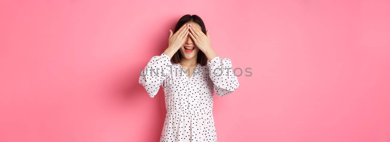 Happy romantic asian girl waiting for surprise, close eyes and smiling amazed, anticipating something, standing over pink background.