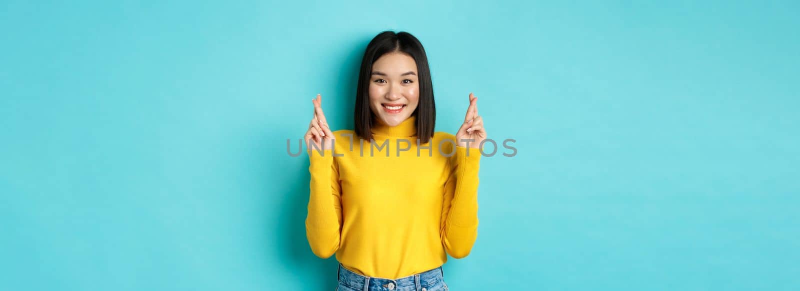 Hopeful and optimistic asian woman cross fingers for good luck, making wish and smiling, looking at camera and expecting something, standing over blue background.