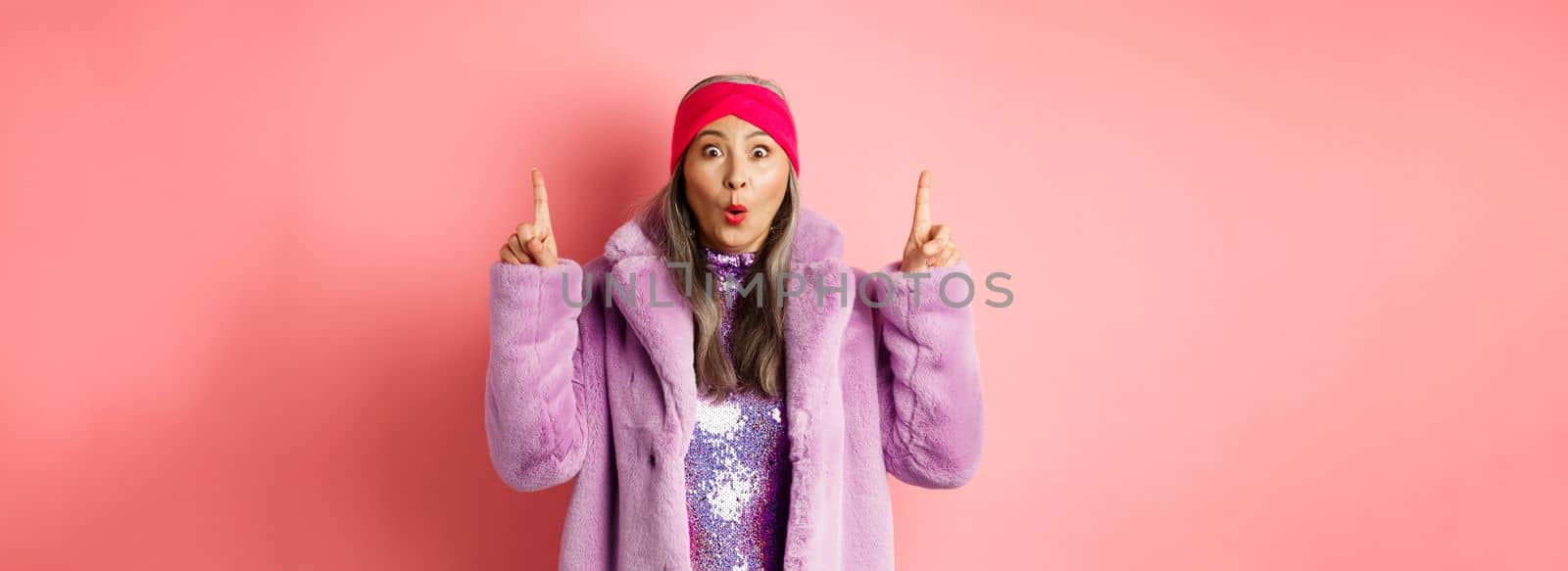 Wow check this out. Amazed asian senior woman pointing fingers up, staring at camera impressed, showing fashion or beauty advertisement, pink background.