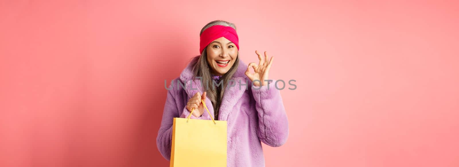 Shopping and fashion. Happy asian senior woman in stylish clothes holding paper bag from store, showing OK sign, recommending shop, pink background.