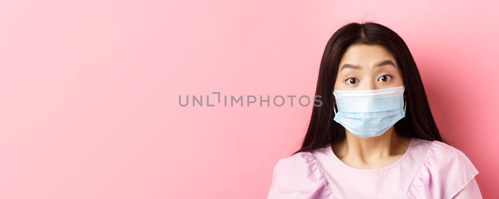 Covid-19 and healthy people concept. Close-up portrait of surprised asian girl in medical mask looking excited at camera, standing against pink background.