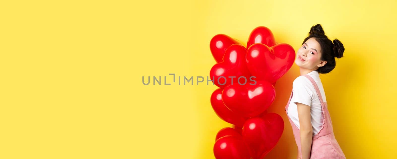 Valentines day and relationship concept. Fashionable asian woman posing near red hearts balloons, surprise for lover on special date, yellow background.