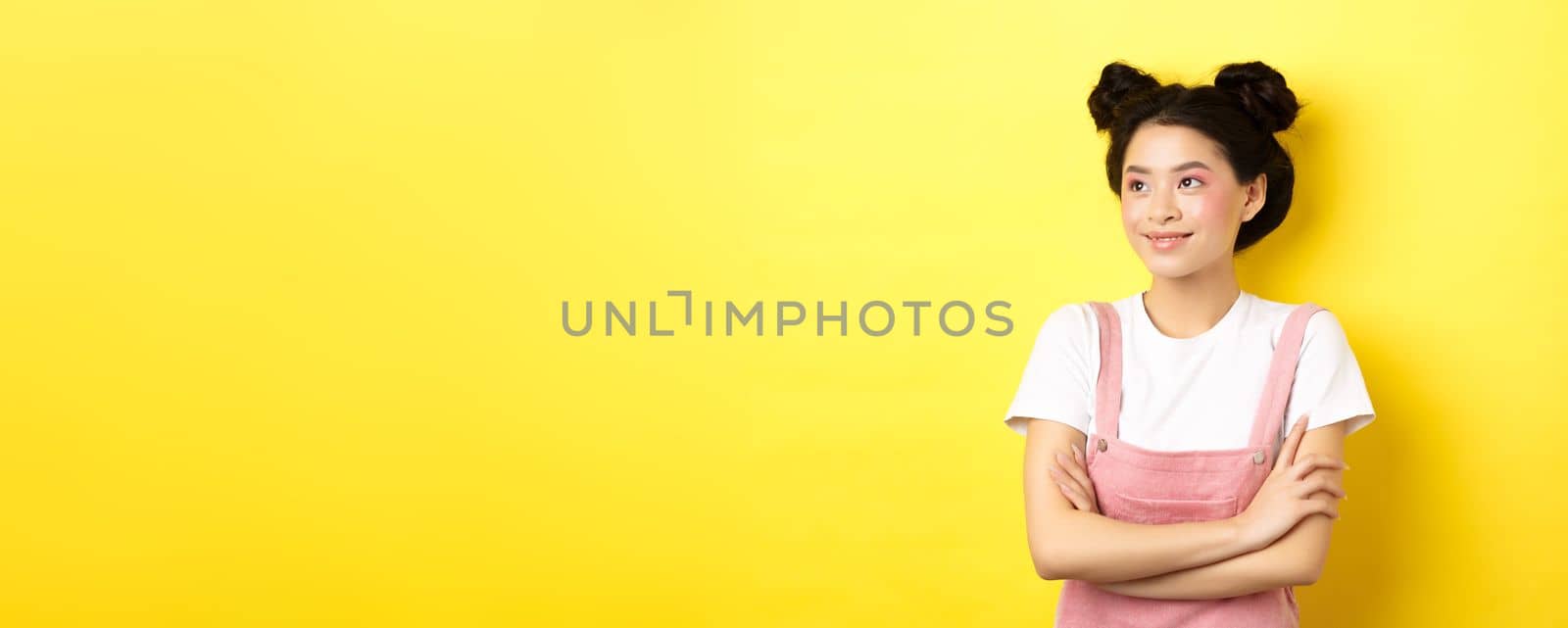 Smiling beautiful asian girl with glamour makeup, looking aside at logo with happy face, standing in summer clothes on yellow background by Benzoix