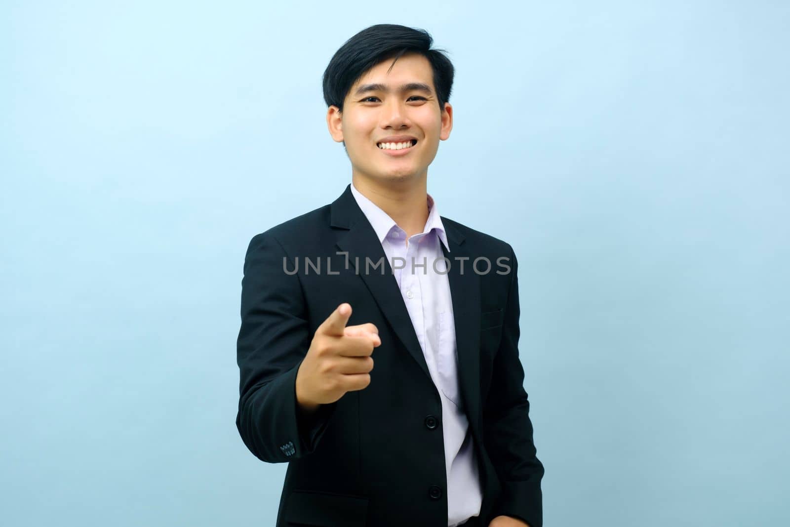 Portriat of asian young smart happy businessman dressed in suit standing straight, smiling, and pointing finger at camera with isolated light blue background. Business concept.