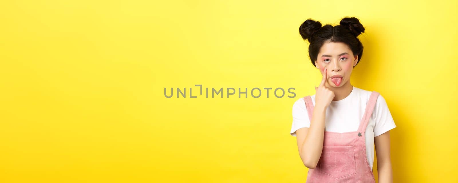 Rude asian girl stretch eyelid and showing tongue, mocking someone, standing on yellow background by Benzoix