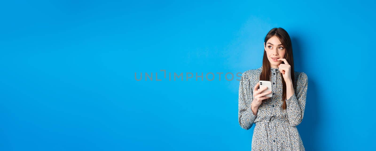 Pensive cute woman thinking how to answer on message, looking aside thoughtful and holding smartphone, standing in dress on blue background by Benzoix
