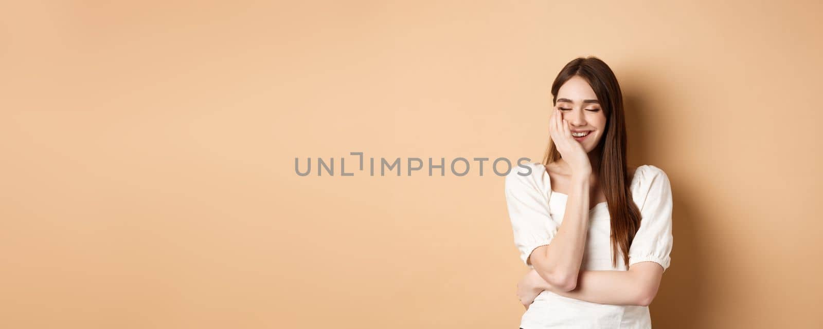 Happy and romantic girl laughing with eyes closed, touching face and feeling joy, standing on beige background by Benzoix