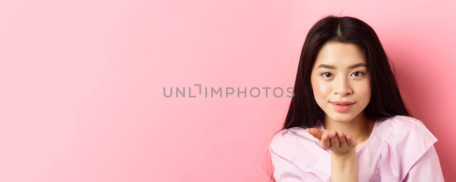 Beautiful asian girl with natural makeup, blowing air kiss at camera with sensual gaze, standing romantic against pink background.