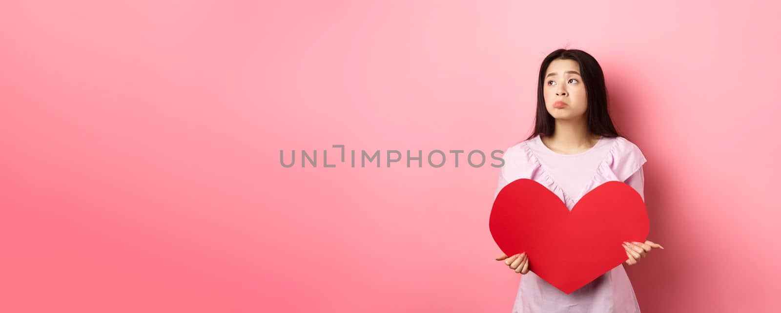 Valentines concept. Lonely teenage asian girl dreaming about love, feeling sad and lonely on lovers day, looking aside with pity, holding big red heart, pink background by Benzoix