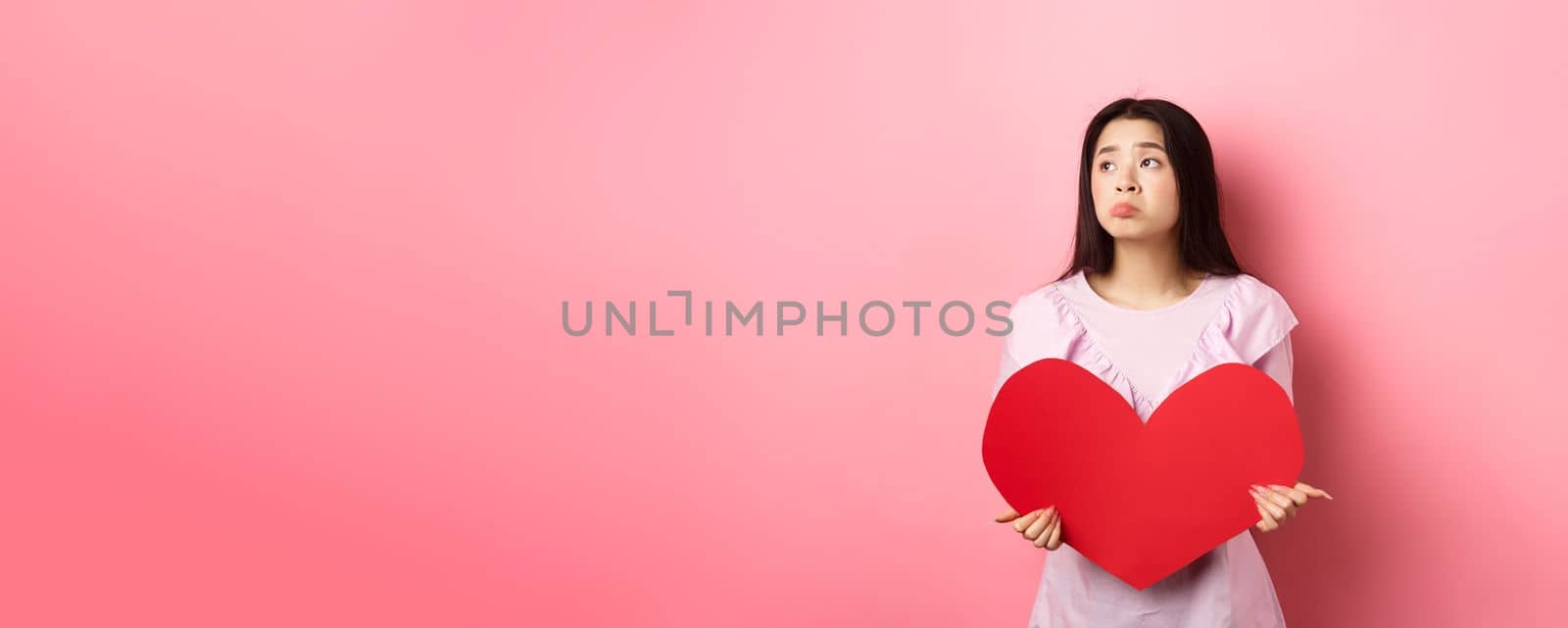 Valentines concept. Lonely teenage asian girl dreaming about love, feeling sad and lonely on lovers day, looking aside with pity, holding big red heart, pink background.