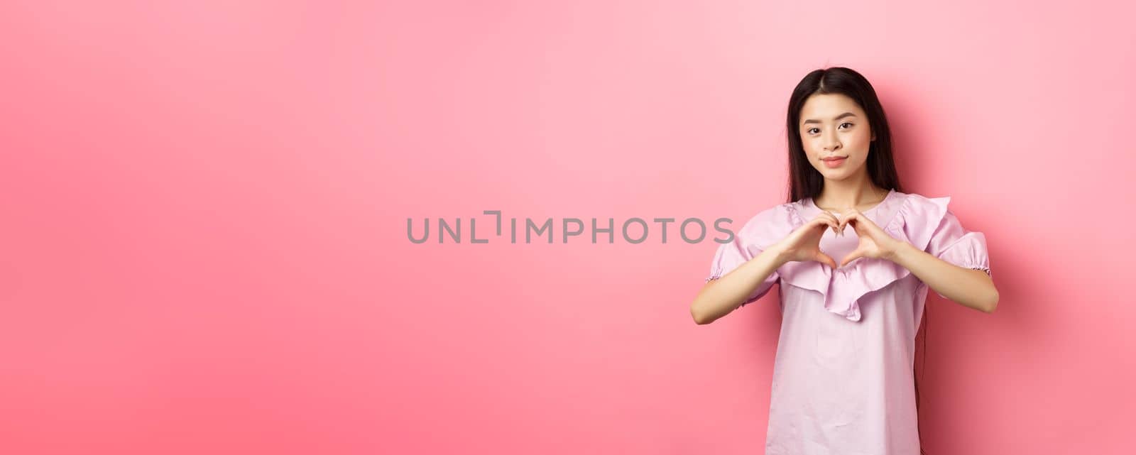 Valentines day concept. Beautiul japanese teen girl show heart gesture and smiling cute, express love and sympathy to lover, standing in romantic dress on pink background by Benzoix