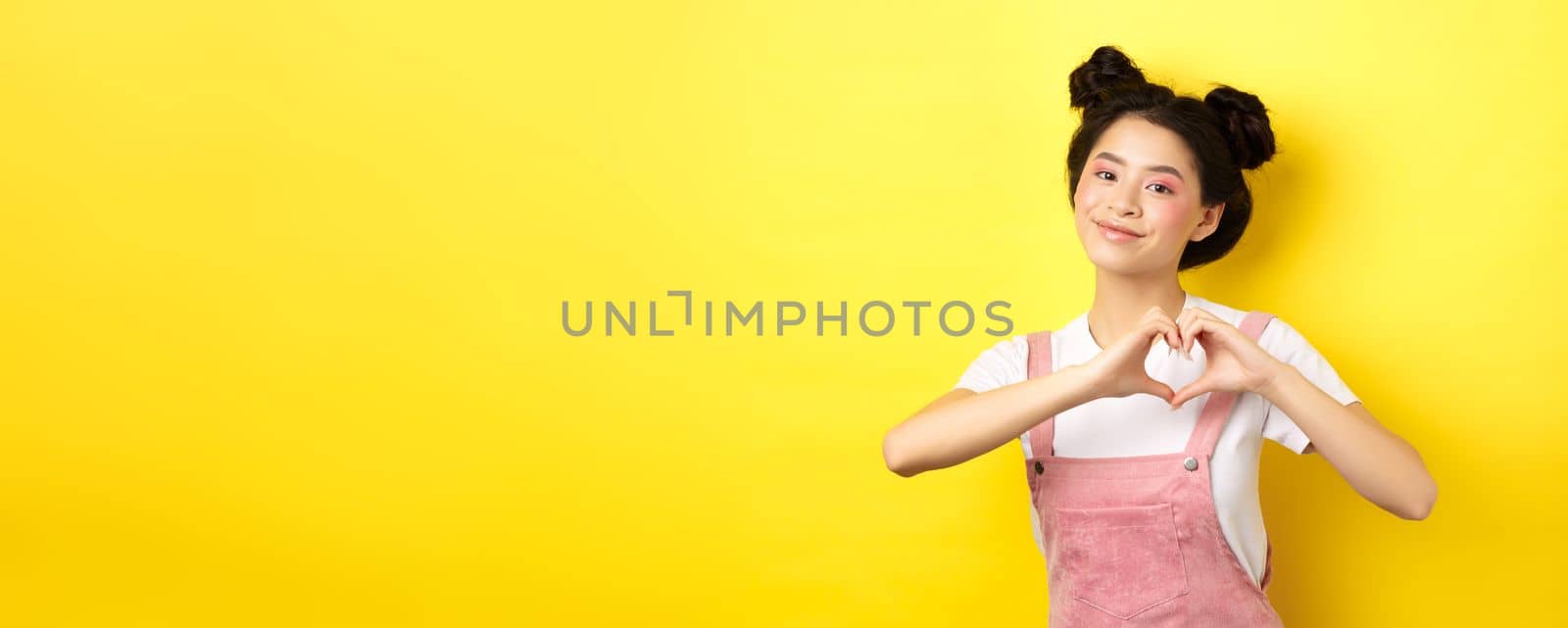 Valentines day. Cute asian girl sending her love, showing heart gesture and smiling at camera romantic, standing on yellow background by Benzoix