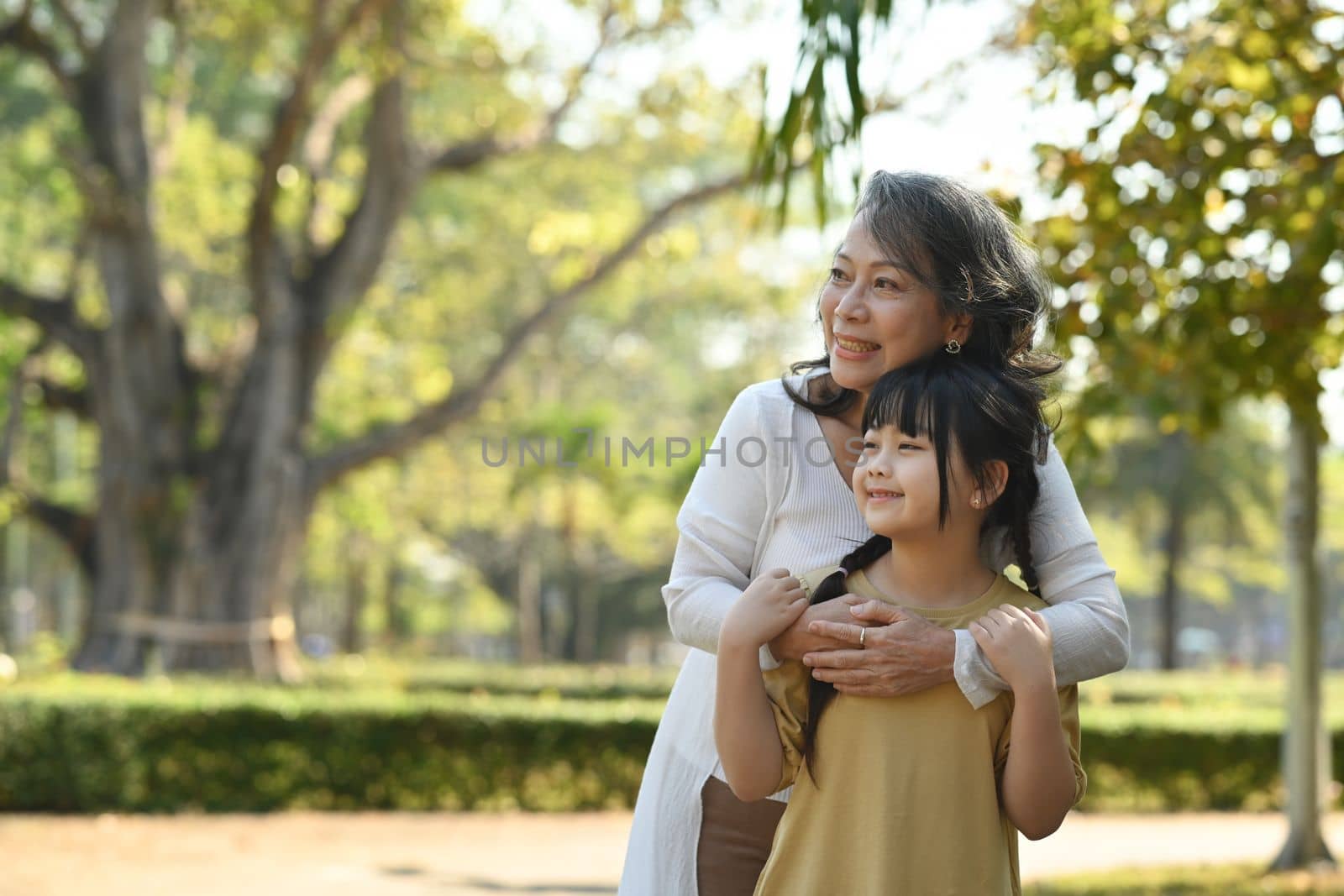 Loving middle aged woman embracing little granddaughter standing outdoor surrounded by beautiful nature view on background.