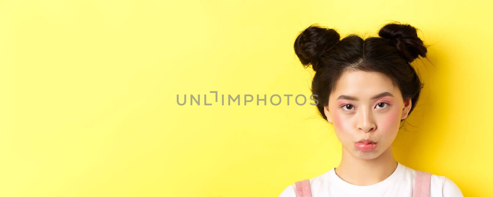 Close up portrait of moody asian girl pouting and looking upset at camera, standing wiht glamour makeup and hairbuns on yellow background.
