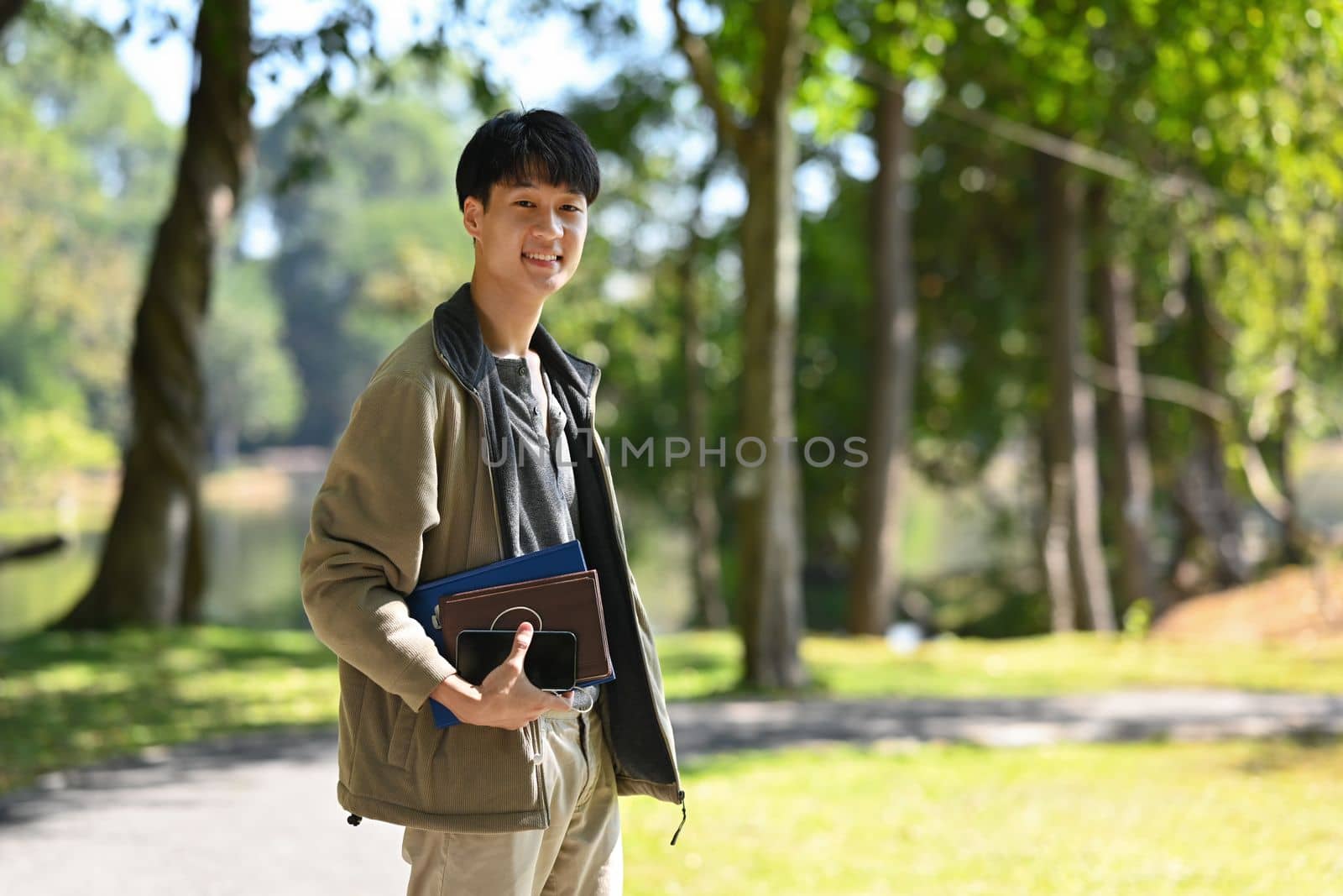 Portrait of young asian man standing near lake with sunny summer park on background. People and recreation in nature.