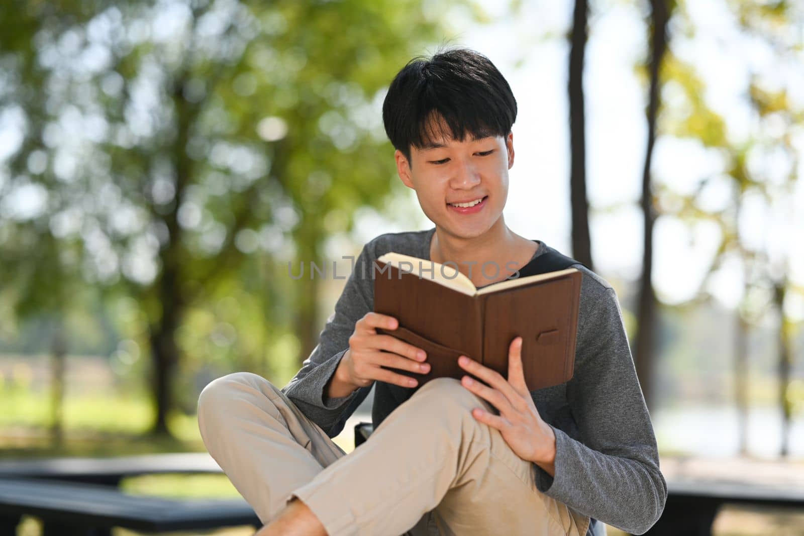 Happy young asian man reading interesting book while sitting bench in sunny summer park by prathanchorruangsak