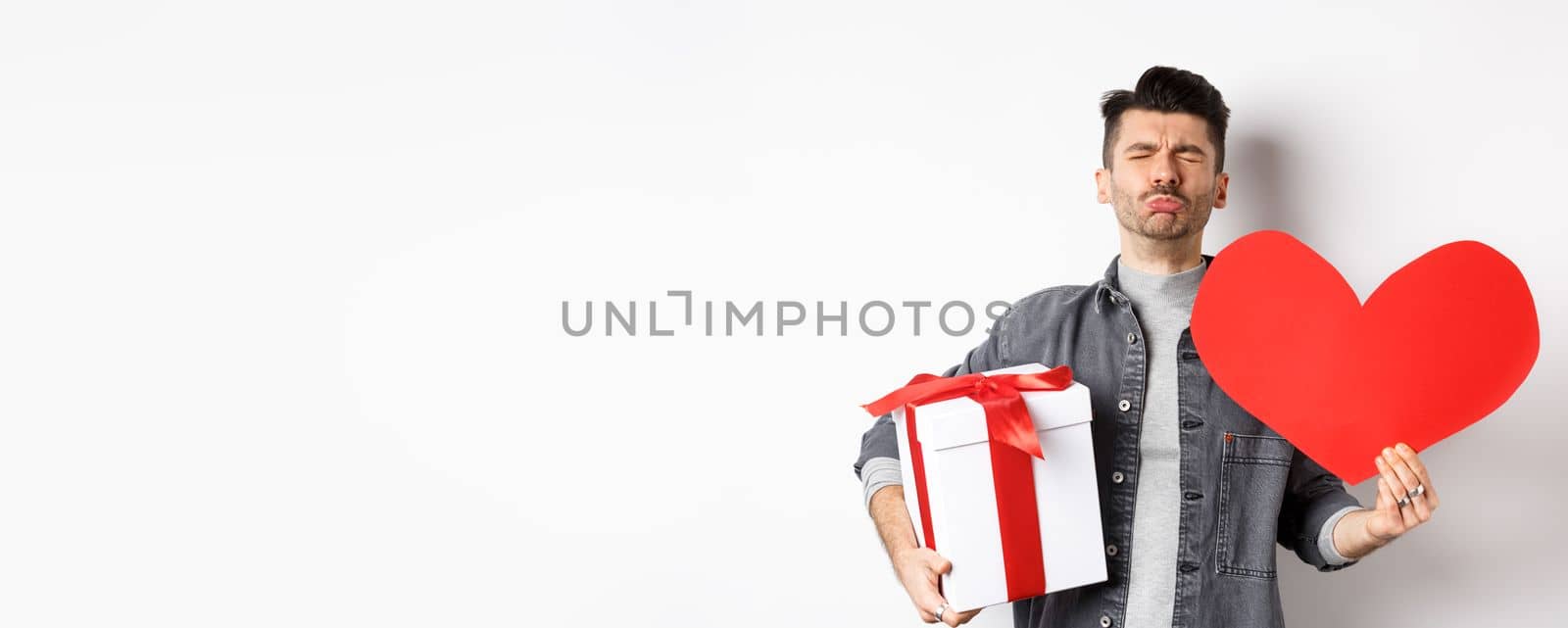 Sad and heartbroken man being rejected, crying and holding red heart with gift box, breakup on Valentines day, white background. Love and relationship concept.