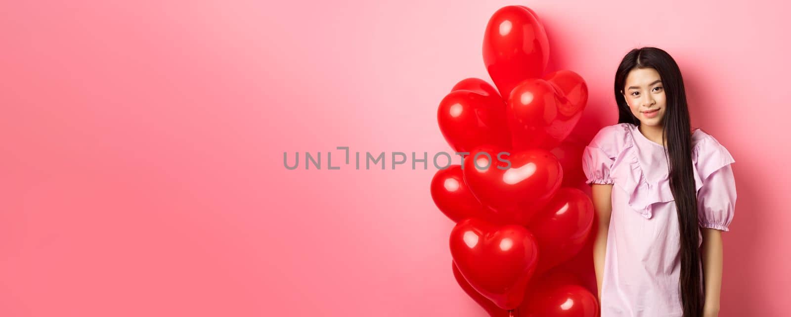 Cute and tender girl looking with love, celebrating valentines and white day with lover, standing near romantic heart balloons on pink background. Relationship concept by Benzoix