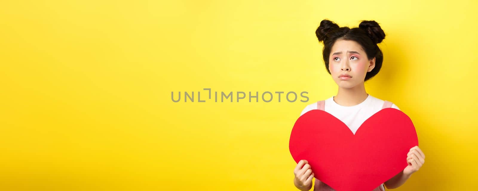 Sad and lonely asian girl standing in make-up with big red heart cutout, left alone on Valentines day, looking left disappointed or upset, standing on yellow background by Benzoix