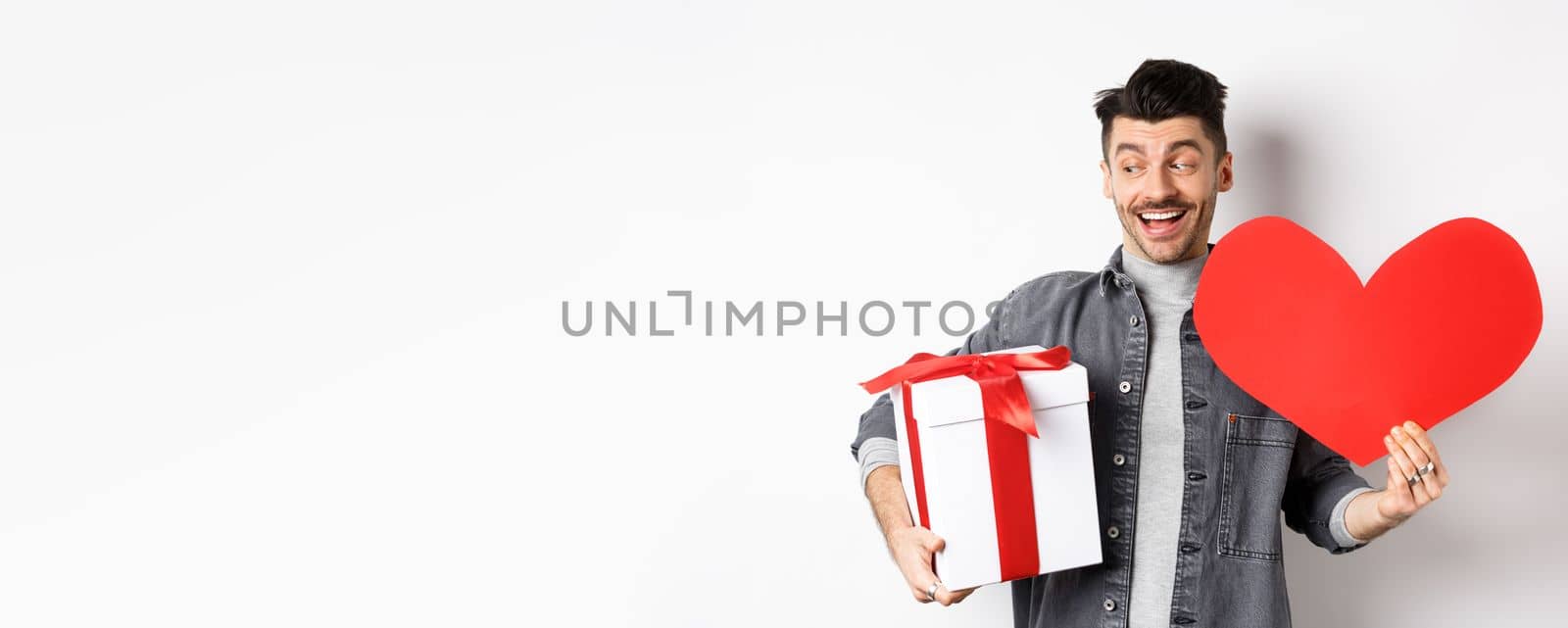 Valentines day and love concept. Handsome man holding gift box and red heart cutout for lover, going on romantic date, smiling excted and looking at empty space, white background by Benzoix