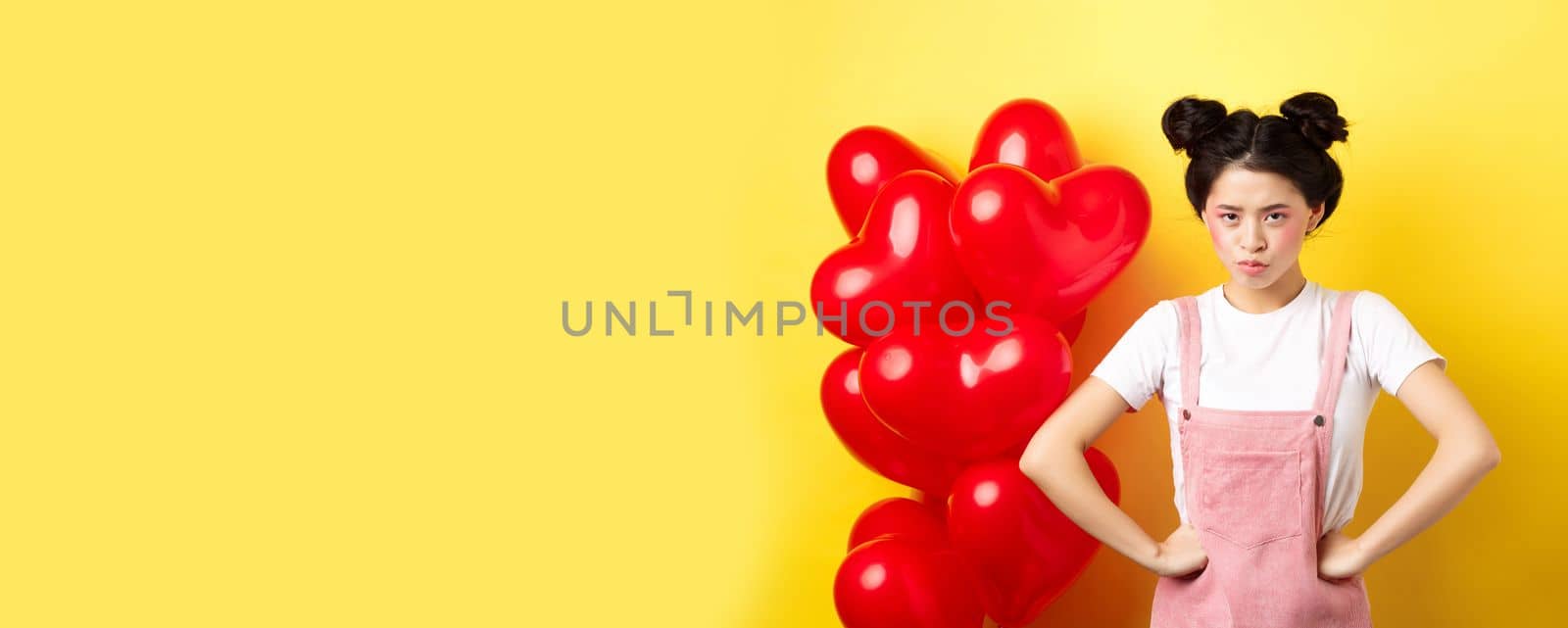 Valentines day and relationship concept. Disappointed asian girlfriend sulking, looking offended at camera, mad at boyfriend, standing near heart balloons, yellow background.