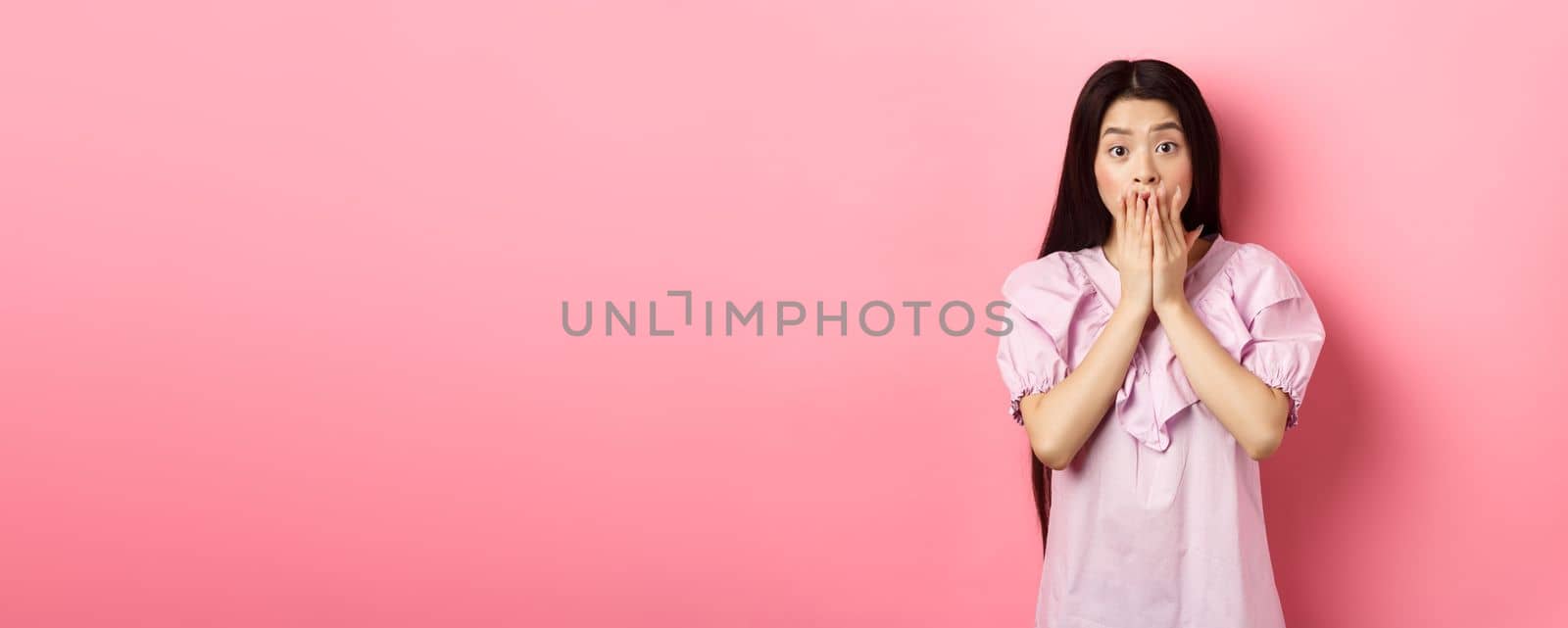 Shocked asian girl with long dark hair, gasping and covering mouth with hands, look at bad news, terrible accident, standing on pink background by Benzoix